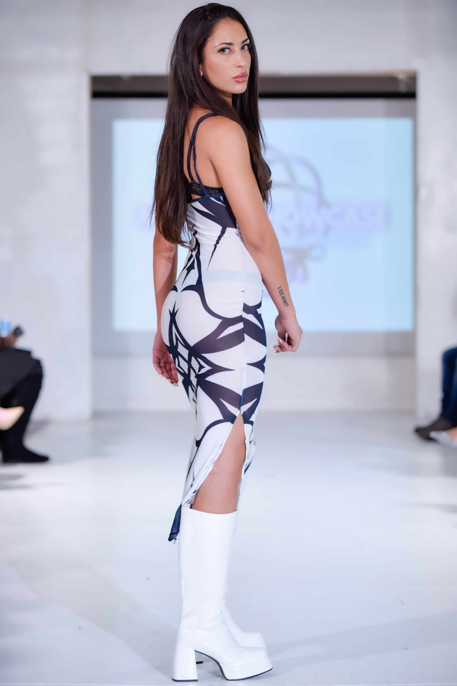 Woman in a patterned dress with a high slit and white boots on a runway.